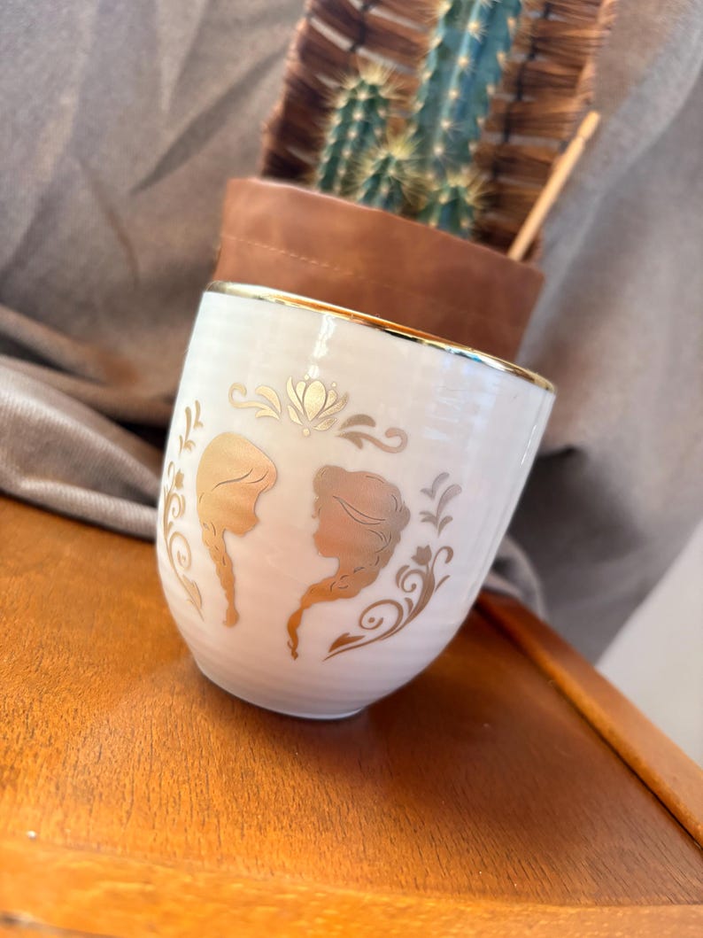 May include: A white ceramic mug with a gold rim and gold silhouette designs. The mug features two female profiles and decorative floral elements. The mug is set on a wooden surface.