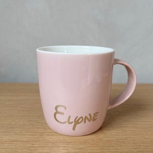 May include: A light pink ceramic mug with a white interior and a curved handle. The exterior features the name Elyne written in a metallic gold script font. The mug has a smooth, glossy finish and a simple, rounded cylindrical shape.