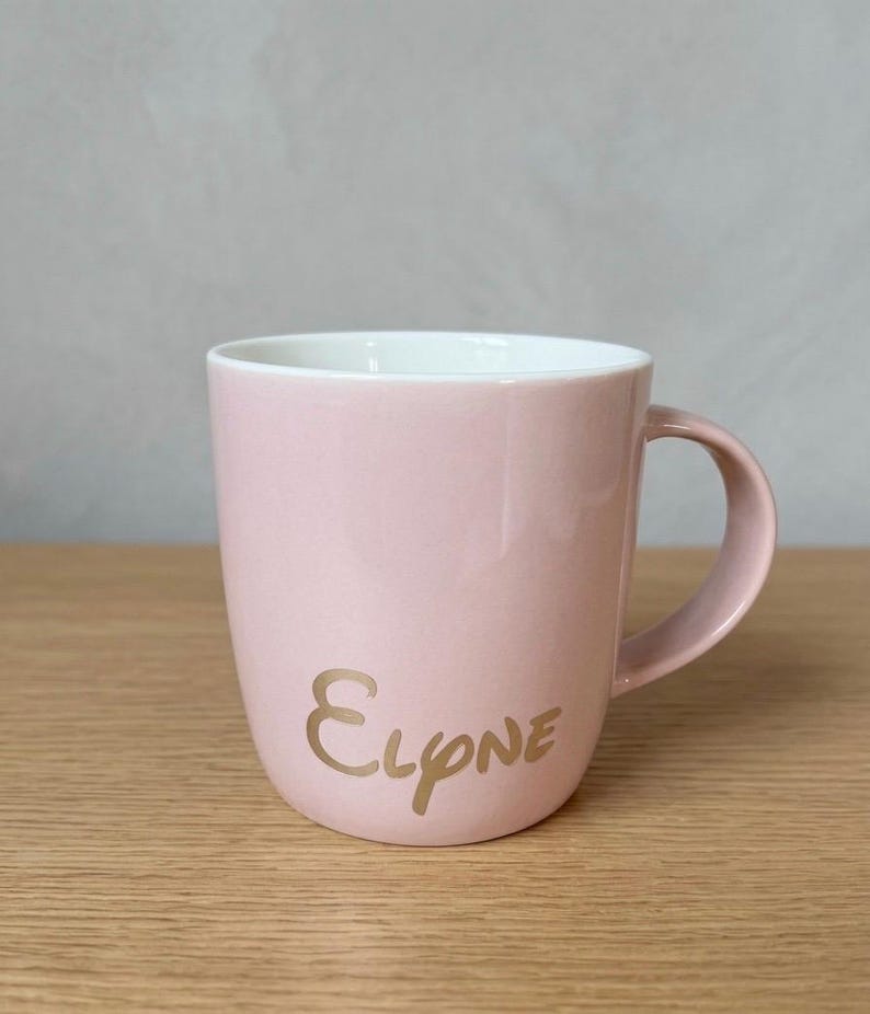 May include: A light pink ceramic mug with a white interior and a curved handle. The exterior features the name Elyne written in a metallic gold script font. The mug has a smooth, glossy finish and a simple, rounded cylindrical shape.