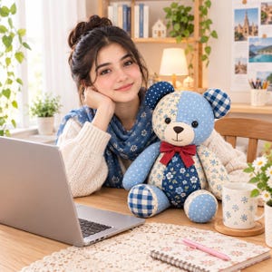 May include: A plush teddy bear constructed from various blue and cream fabric patches featuring floral, polka dot, and gingham patterns. The bear has dark button eyes, a stitched nose and mouth, and a solid dark red ribbon bow tied around its neck.