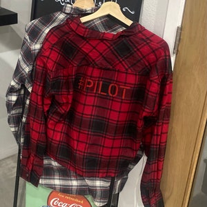 May include: Red and black plaid flannel shirt with long sleeves and a button-up front. The shirt has the text "#PILOT" printed on the back.