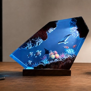 May include: A resin lamp depicting an underwater scene with a whale, sea turtles, and coral. The lamp emits a blue light and rests on a wooden base. The lamp is shaped like a slanted hexagon.