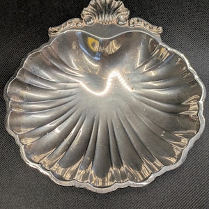 May include: A silver-toned metal dish shaped like a scallop shell. The dish features a polished, reflective surface with deep, radiating fluted grooves and a decorative, scalloped edge detail at the top center, creating a classic, shell-like form.