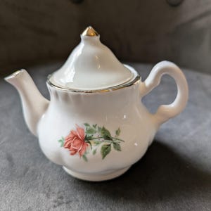 May include: A miniature white porcelain teapot with gold trim and a pink rose design. The teapot has a curved handle and a lid with a gold finial. A delicate item, perfect for serving tea. The teapot measures approximately 15 cm in length.