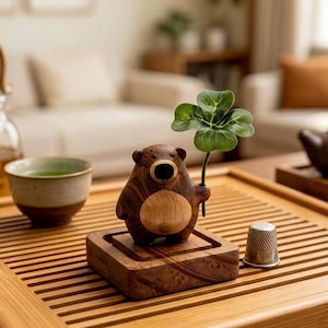 May include: A small wooden bear figurine with a dark brown grain and a lighter circular belly, holding a green four-leaf clover. The bear stands on a square wooden base next to a small silver metal thimble on a slatted wooden tray.