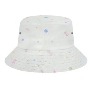 May include: White bucket hat with a pastel floral and bow pattern. The hat features a wide brim with small perforations and two black eyelets on the sides. The design includes pink, blue, and yellow accents.