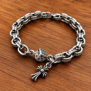 May include: A silver chain link bracelet featuring circular links engraved with small cross symbols. The bracelet includes a dangling charm shaped like a six-sided die and a separate dangling charm shaped like a decorative cross pendant.