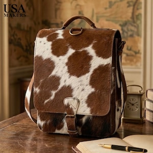 May include: A brown and white cowhide backpack featuring a top handle, a front flap with a buckle closure, and a metal D-ring at the top. The bag has a textured fur finish with a spotted pattern. The text USA MAKERS is visible in the top left corner.