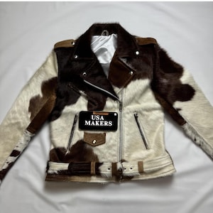 May include: A cowhide leather motorcycle jacket featuring a brown and white spotted pattern, a silver-tone asymmetrical front zip, a small flap pocket with a snap button, and a buckled waist belt. A black tag displays the text USA MAKERS.