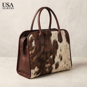 May include: A structured handbag featuring a brown and white cowhide panel on the front, complemented by smooth dark brown leather sides, handles, and trim. The bag includes a top zipper closure with metallic hardware and the text USA MAKERS.