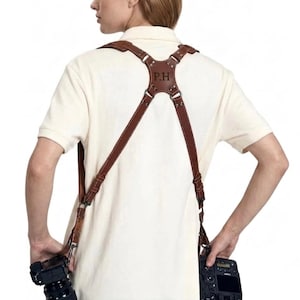 May include: A brown leather dual camera harness featuring a central back plate embossed with the letters P.H. The harness includes adjustable straps, metal hardware, and clips attached to two black digital cameras held at the sides.
