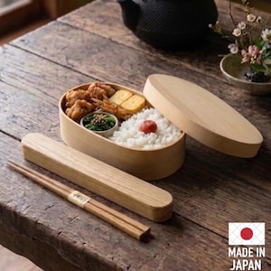 May include: An oval wooden bento box with a matching lid, filled with white rice topped with a pickled plum, fried chicken, rolled omelet, and seasoned greens. A rectangular wooden chopstick case and a pair of wooden chopsticks are included.