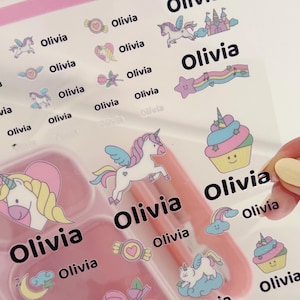 May include: A clear sheet of adhesive labels featuring the name Olivia in black text alongside colourful illustrations of unicorns, castles, cupcakes, rainbows, stars, and hearts. The top of the sheet is labelled with the word UNICORN in pink.
