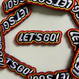 Könnte beinhalten: Gestickte Aufnäher mit dem Text "LET'S GO!" in Weiß, umrandet von Gelb und Schwarz, mit rotem Rand. Die Aufnäher sind auf einer hellen Oberfläche verstreut. Die Aufnäher sind wahrscheinlich für Kleidung oder Accessoires.