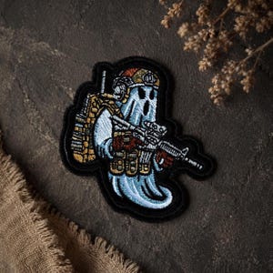 Può includere: Una toppa ricamata raffigurante un fantasma azzurro che indossa un casco tattico beige, uno zaino e un gilet abbinati. Il fantasma tiene in mano un fucile nero, realizzato con cuciture dettagliate e rifinito con un bordo nero.