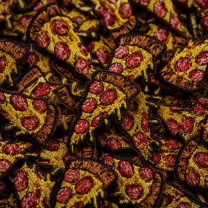 Pode incluir: Uma coleção de emblemas bordados em forma de fatia de pizza com queijo derretido amarelo e coberturas de pepperoni vermelho. Cada emblema tem uma borda costurada em marrom escuro e retrata uma fatia triangular com detalhes texturizados.
