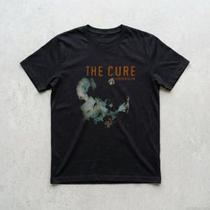 Könnte beinhalten: Schwarzes T-Shirt mit dem Bandnamen "THE CURE" in braunem Text über einem Gesicht mit floralem Design. Das Wort "DISINTEGRATION" steht unter dem Bandnamen. Ein Artikel zum Thema Musik.