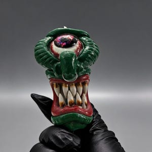 May include: A green, red, and white monster candle with a large eye, sharp teeth, and a nose ring. The candle is held by a gloved hand against a gray background. The candle has a unique, artistic design.