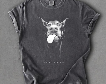 Puede incluir: Camiseta color pimienta con un gráfico en blanco y negro de un perro Doberman sacando la lengua. La palabra "DOBERMAN" está impresa debajo de la imagen del perro. La camiseta tiene las mangas enrolladas.