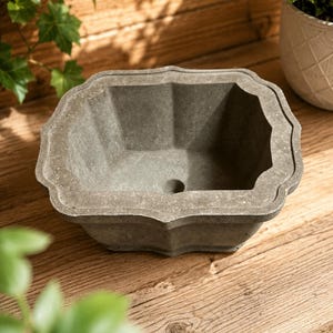 Puede incluir: Una jardinera rectangular gris con un borde festoneado. La jardinera tiene un diseño geométrico y un orificio de drenaje. La jardinera está hecha de un material similar al hormigón y es adecuada para uso en exteriores. La jardinera está sobre una superficie de madera.