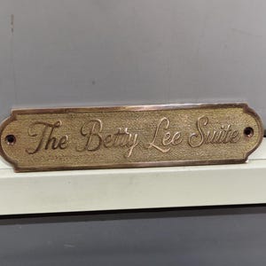 May include: A rectangular brass door plaque with rounded ends and two mounting holes. The metal surface has a textured finish and features the engraved script text "The Betty Lee Suite" centered across the front of the plate in dark lettering.
