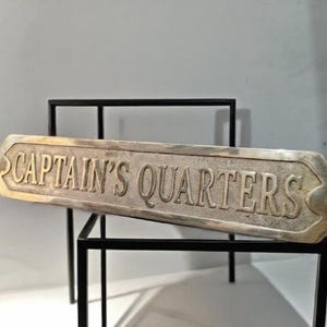 Pode incluir: Uma placa de metal retangular com extremidades pontiagudas apresentando o texto em relevo "CAPTAIN'S QUARTERS" em uma fonte serifada. A placa tem um acabamento metálico desgastado com um fundo texturizado e dois pequenos orifícios.