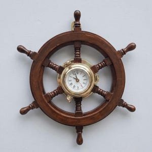 Puede incluir: Un reloj de pared con forma de timón de barco de madera con un acabado marrón oscuro y seis radios. El centro presenta una carcasa redonda de latón con una esfera blanca, manecillas negras y números romanos negros para las horas.