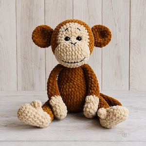 May include: A plush monkey toy crafted from soft brown and cream yarn. The monkey features a textured, crocheted appearance with large round ears, a light-coloured face, black button eyes, and a stitched mouth, sitting in an upright position.