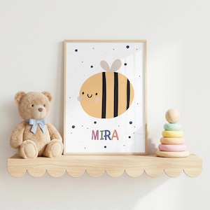 Puede incluir: Una lámina artística enmarcada con una abeja de dibujos animados con cuerpo amarillo, rayas negras y cara sonriente, rodeada de pequeños puntos azul oscuro. El nombre MIRA está impreso en letras de colores bajo la abeja sobre fondo blanco.