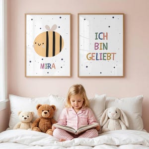 Puede incluir: Dos láminas decorativas rectangulares en marcos de madera clara. La lámina de la izquierda muestra una ilustración de una abeja amarilla y negra con el nombre MIRA debajo. La de la derecha muestra el texto colorido ICH BIN GELIEBT. Ambas tienen fondo blanco con puntos azul oscuro.