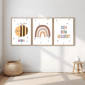 Puede incluir: Tres cuadros decorativos de pared con una abeja de dibujos animados con el nombre MIRA, un arcoíris en tonos marrones y el texto ICH BIN GELIEBT. Cada lámina blanca está decorada con pequeños puntos azul oscuro y un marco de madera clara.