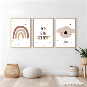 Puede incluir: Tres láminas rectangulares en marcos de madera clara. La lámina de la izquierda muestra un arcoíris marrón y beige. La central presenta el texto "ICH BIN GELIEBT" en letras multicolores. La de la derecha muestra una cara de koala y "LINA".