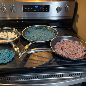 May include: Three metal frying pans with silver handles, two containing round crocheted doilies in teal and mauve, and one covered pan containing a cream-colored doily. A separate teal crocheted doily sits on the black stovetop surface.