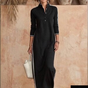 May include: A black maxi dress with white stripes down the sides and sleeves. The dress has a high collar and a button-down front. A woman is holding a white woven tote bag and wearing sandals. The background is a neutral color with decorative pottery.