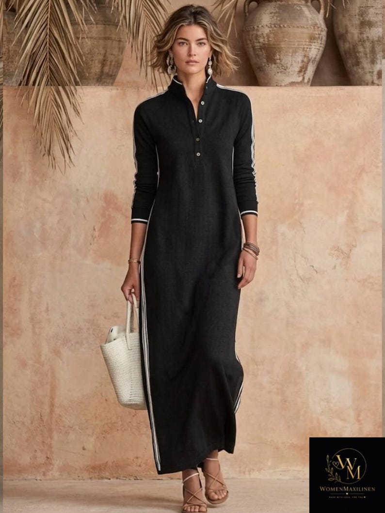 May include: A black maxi dress with white stripes down the sides and sleeves. The dress has a high collar and a button-down front. A woman is holding a white woven tote bag and wearing sandals. The background is a neutral color with decorative pottery.