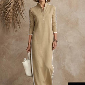 May include: A long, tan linen dress with a mandarin collar and white trim. The dress features white stripes down the sides and a button-up front. The model is holding a white tote bag. The logo "WOMENMAXILINEN" is in the bottom right corner.