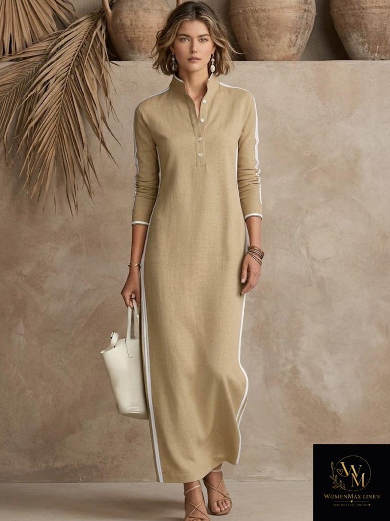 May include: A long, tan linen dress with a mandarin collar and white trim. The dress features white stripes down the sides and a button-up front. The model is holding a white tote bag. The logo "WOMENMAXILINEN" is in the bottom right corner.