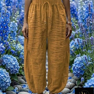 May include: Mustard-colored linen pants with a drawstring waist and pockets. The pants have a relaxed fit and are gathered at the ankles. The image is set against a backdrop of blue flowers and a stone path. The pants are part of the Women Maxilinen collection.