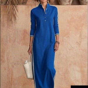 May include: A long, blue dress with white stripes down the sides and sleeves. The dress has a mandarin collar and a button-down front. A woman is holding a white tote bag. The background features neutral tones and decorative pottery.