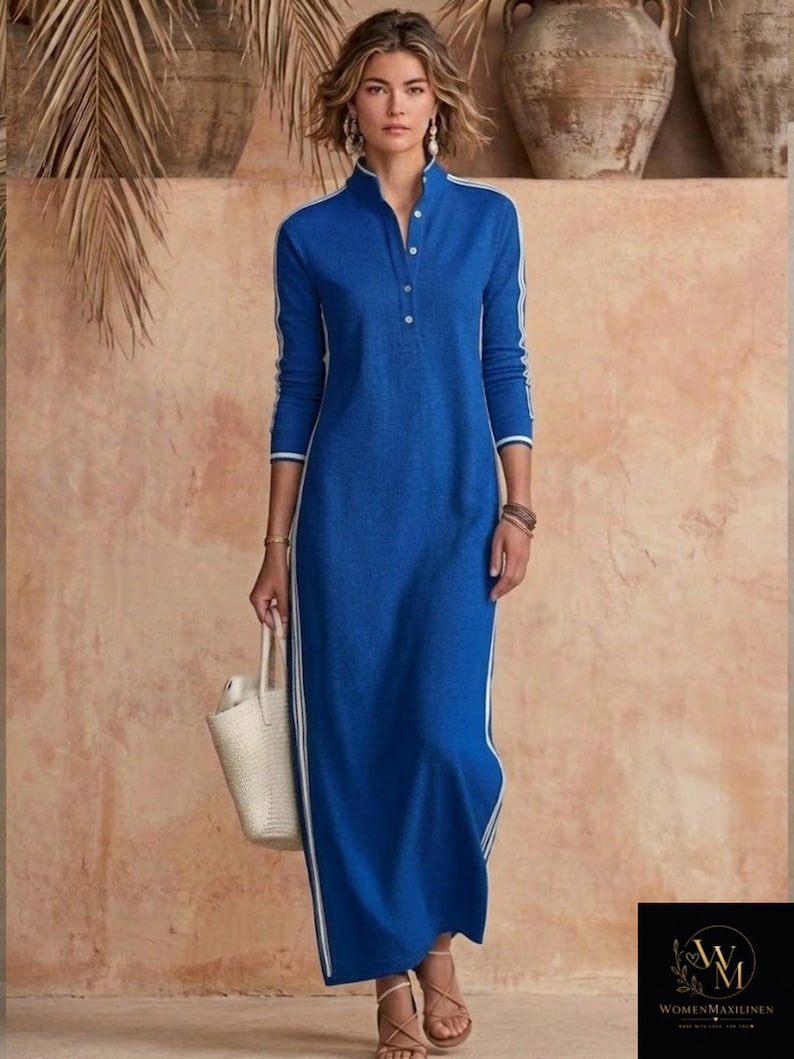 May include: A long, blue dress with white stripes down the sides and sleeves. The dress has a mandarin collar and a button-down front. A woman is holding a white tote bag. The background features neutral tones and decorative pottery.