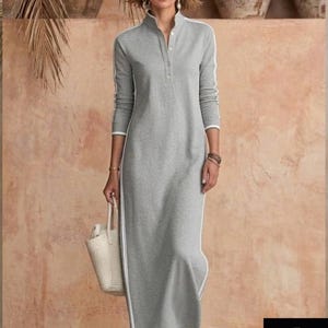May include: A gray maxi dress with white stripes down the sides and sleeves. The dress has a high collar and a button-down front. A woman is wearing the dress and carrying a white tote bag. The background is a neutral color.