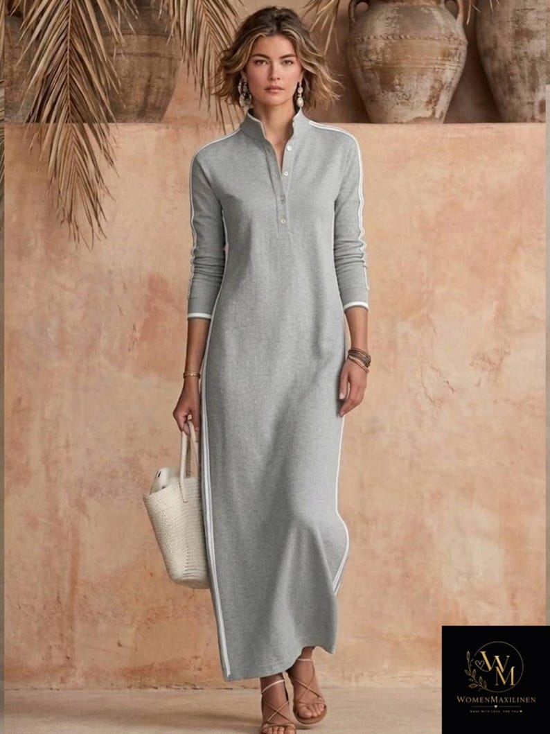 May include: A gray maxi dress with white stripes down the sides and sleeves. The dress has a high collar and a button-down front. A woman is wearing the dress and carrying a white tote bag. The background is a neutral color.