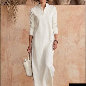 May include: A long, off-white linen dress with a mandarin collar and long sleeves. The dress features white trim along the sides and sleeves. A woman is wearing the dress and carrying a woven tote bag. The background is a warm, earthy tone.