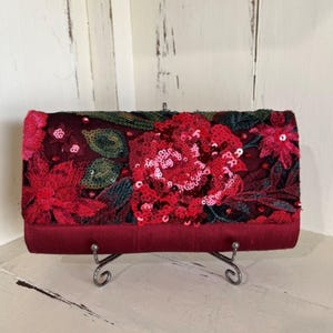 May include: A rectangular clutch bag in deep burgundy satin featuring an ornate flap adorned with intricate floral embroidery, dark green leaf stitching, and vibrant red sequins that create a textured, shimmering surface across the front.