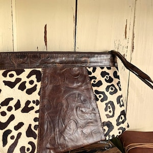 May include: A rectangular clutch bag featuring a central panel of dark brown embossed leather with a swirling pattern, flanked by two side panels of cream-coloured hair-on hide with a black leopard print. The bag includes a matching leather wrist strap.