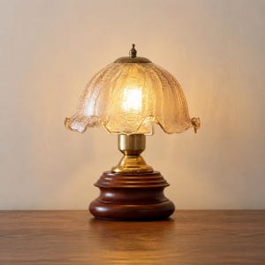 May include: A table lamp featuring a dark brown wooden base, a polished brass neck, and a translucent, amber-tinted glass shade with a ruffled, scalloped edge. The lamp emits a warm, soft glow from the light bulb visible inside the textured glass.