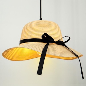 May include: A pendant light fitting shaped like a straw hat with a wide brim and a rounded crown. The hat is light beige with a woven texture and features a black ribbon tied in a bow around the base of the crown. A single light bulb is visible.