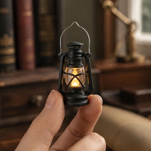 Puede incluir: Una linterna negra en miniatura con un globo de cristal transparente y un asa de alambre plateado fino. La linterna cuenta con una pequeña luz LED de color amarillo cálido en su interior, que imita el aspecto de una lámpara de aceite tradicional.