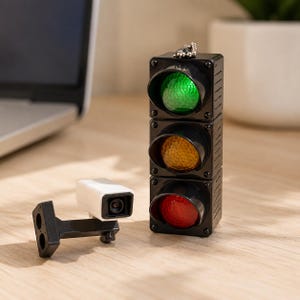 Könnte beinhalten: Ein schwarzer Ampel-Schlüsselanhänger aus Kunststoff mit drei runden Linsen in Grün, Gelb und Rot sowie einer kleinen Metallkugelkette. Daneben befindet sich ein weiß-schwarzes Überwachungskameramodell mit einer schwarzen Halterung.