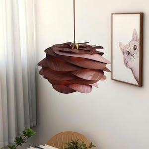 May include: A spherical pendant light fitting constructed from overlapping dark brown wooden petal-shaped slats. The lamp hangs from a thin brown cord attached to a metallic gold-toned fitting at the top, creating a layered, organic, and rounded form.